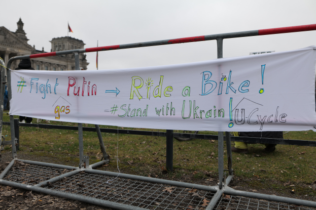 Eine Gruppe von Menschen hält ein Banner mit der Aufschrift "Kämpfe gegen Putin, fahr Rad, steh mit der ukrainischen U-Cycle" und steht auf dem Boden vor einem Metallzaun, trockenen Blättern, einem Gebäude mit Fenstern, einem Fahnenmast mit einer Flagge und einem bewölkten Himmel.