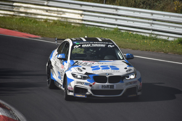 BMW M4 DTM Rennauto auf einer Rennstrecke mit sichtbaren Rädern, umgeben von Gras, einem Zaun und Pflanzen.