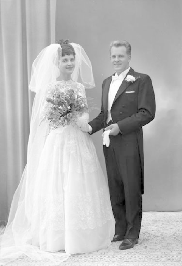 Schwarzes und weißes Foto eines Mannes und einer Frau, die nebeneinander auf ihrer Hochzeit stehen, die Frau in einem weißen Kleid mit einem Strauß, der Mann neben ihr mit einem Lächeln, mit einem Vorhang und einer Wand im Hintergrund.