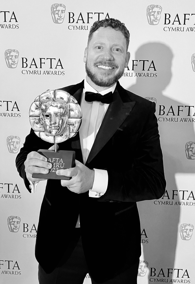 Ein Mann in einem Smoking, der eine BAFTA-Trophäe hält und lächelt, mit einer Hintergrundtafel, auf der "BAFTA Awards 2018" steht.