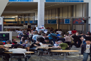 Ein belebter Flughafen-Foyer mit wartenden Menschen, einige stehen und andere arbeiten, Richtungsschilder an der rechten Wand und darüber sowie Lampen, die von der Decke hängen.