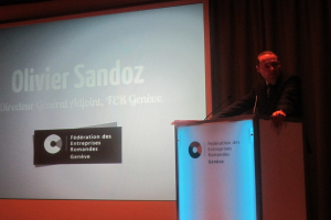 Eine Person steht auf einem Podium und hält eine Rede, vor ihr befinden sich zwei Mikrofone und ein großer Bildschirm.