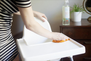 Eine Frau in einem schwarzen und weißen Kleid wischt einen Tisch ab, mit einem Schrank rechts daneben, der eine Flasche, einen Topf und eine Weltkugel enthält.