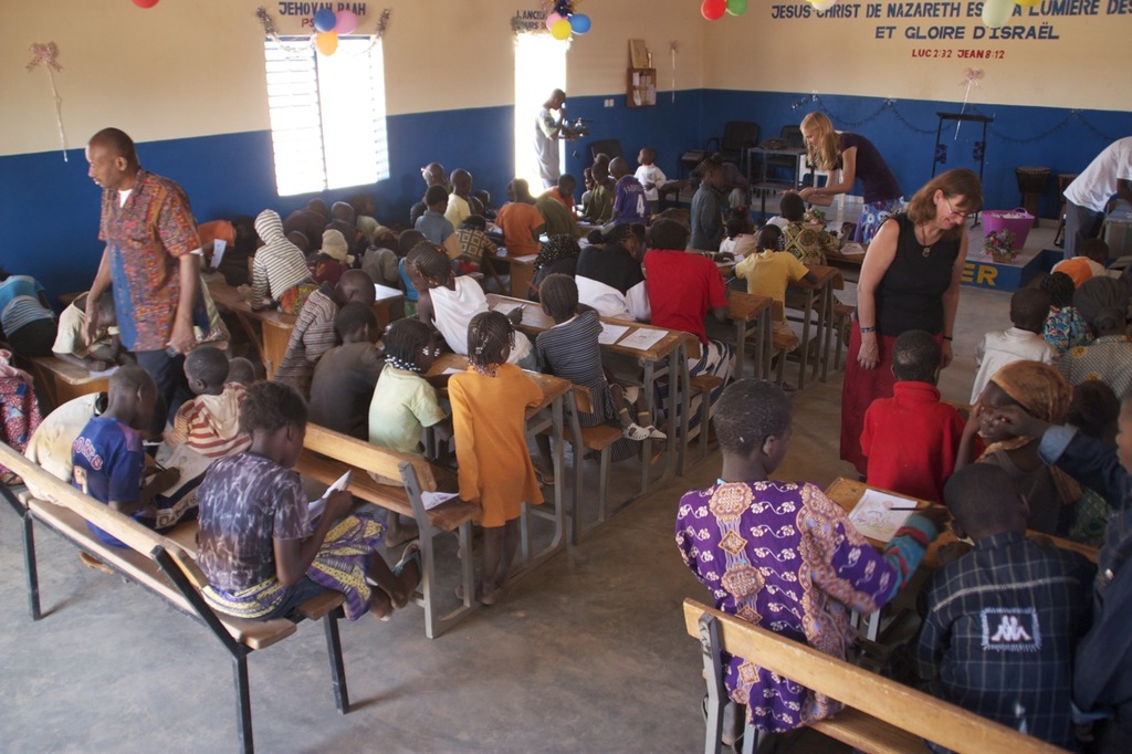 Kinder auf Bänken im Vordergrund, mit Erwachsenen, die sie führen, und einem Hintergrund mit Stühlen, einer Tür, einem Fenster, einem Tisch, Ballons und Text oben.