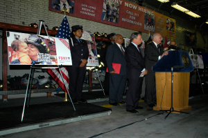 Eine Gruppe von Menschen steht in der Nähe eines Podiums mit Mikrofonen, umgeben von Schildern auf Ständern, Fahnen und Schildern an der Wand, alles unter hellen Scheinwerfern.