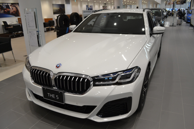 Weißer BMW 5er in einem modernen Showroom mit schwarzer Schlafzimmer-Möblierung, Postern an den Wänden und Deckenbeleuchtung, die eine gemütliche Atmosphäre schafft.