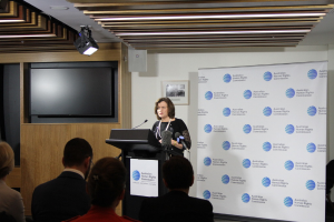 Eine Frau steht auf einer Bühne und spricht in ein Mikrofon, vor ihr sitzt ein Publikum, an der Wand hinter ihr hängt ein Banner, auf dem Holzwand steht ein Fernseher und an der Decke leuchten Lampen.